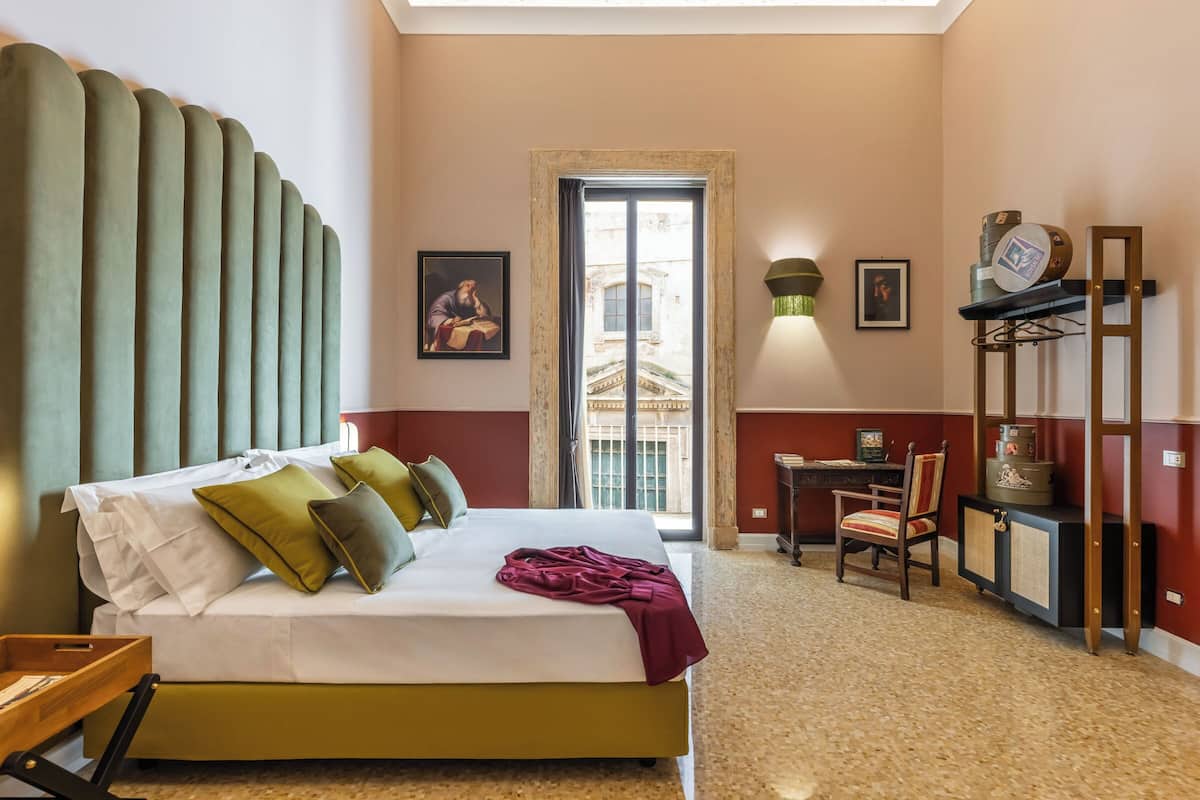Quarto de hotel com cama, escrivaninha, cadeira, janela e porta.