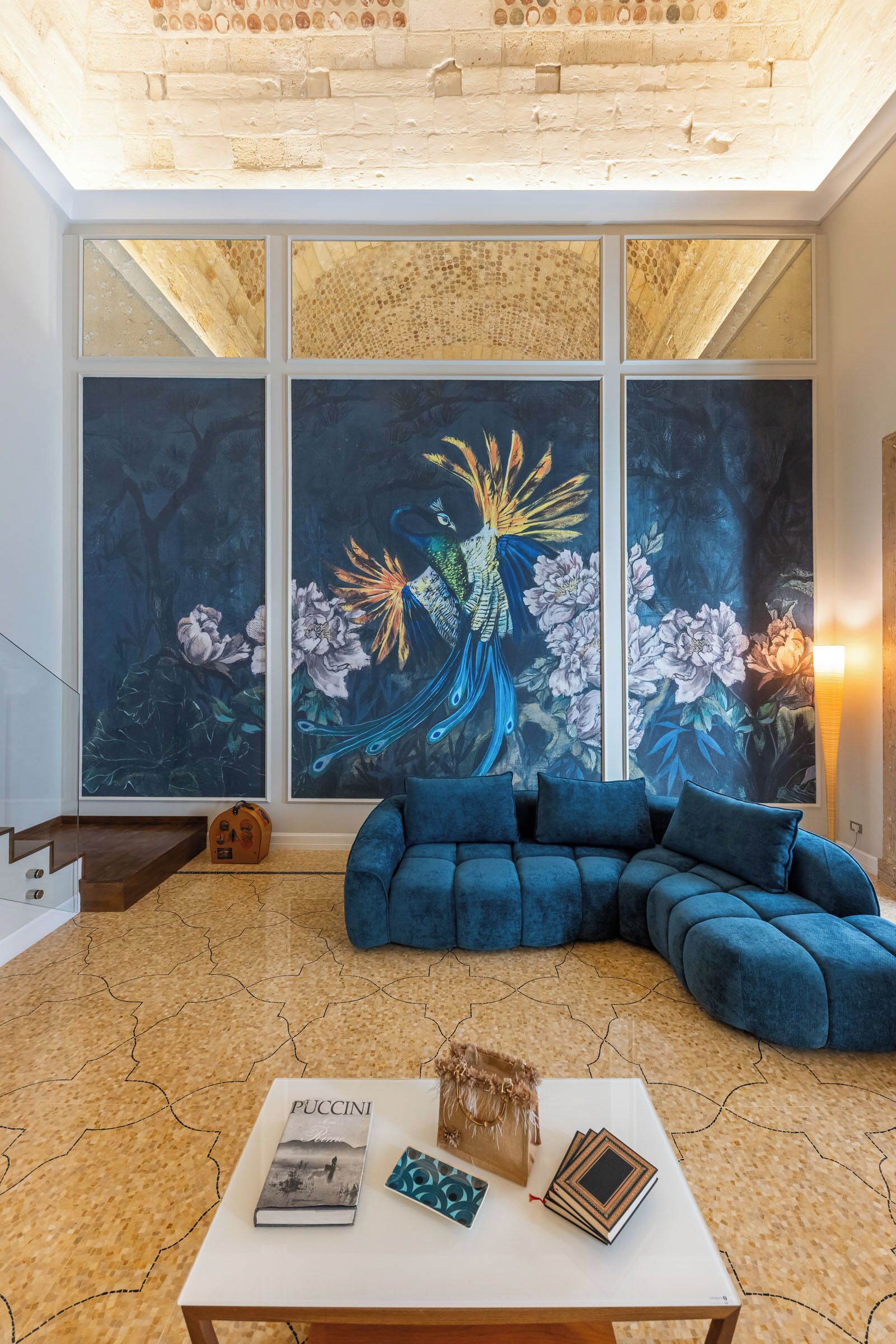 A modern living room with a blue sectional sofa, a white coffee table with books and a decorative item, and a large window with a mural of a peacock.