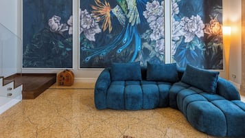 Un moderno salón con un sofá modular azul, una mesa de centro blanca con libros y un objeto decorativo, y un amplio ventanal con un mural de un pavo real.