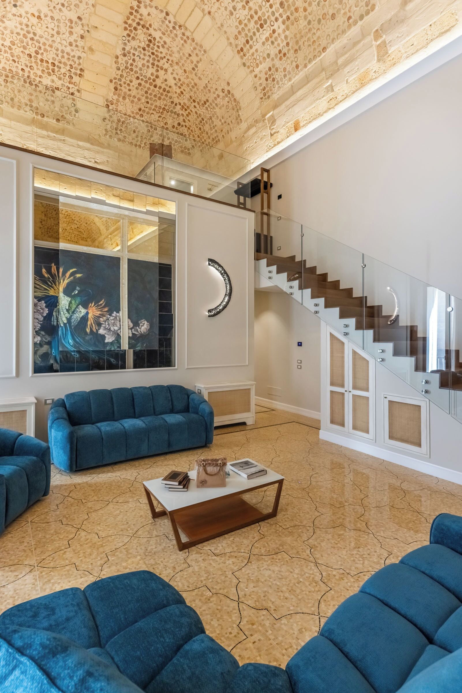 Un espacio habitable amplio con techo alto, un ventanal con vista decorativa, una escalera con barandilla de vidrio y mobiliario tapizado en azul.