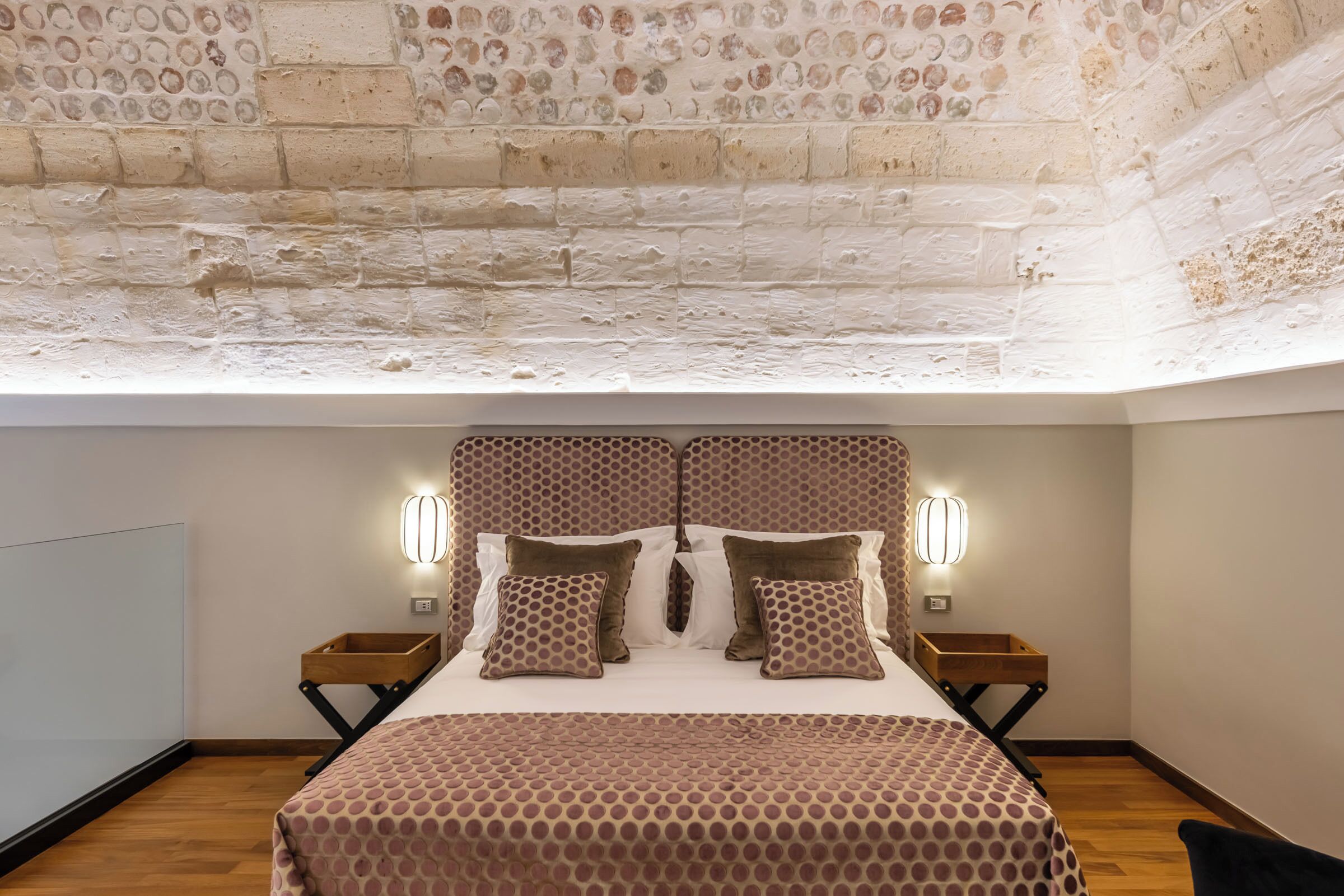 Um quarto com teto em padrão de tijolos, cama com roupa de cama estampada, duas mesas de cabeceira e luminárias fixadas na parede.