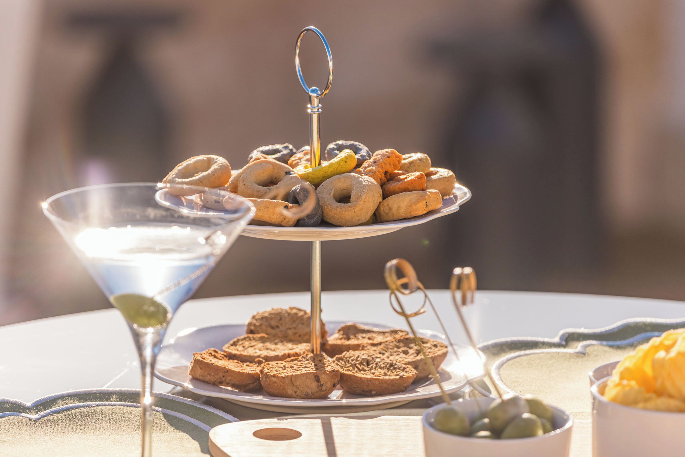 A three-tiered tray of assorted pastries and bread, a martini glass, and a small bowl of olives on a table.