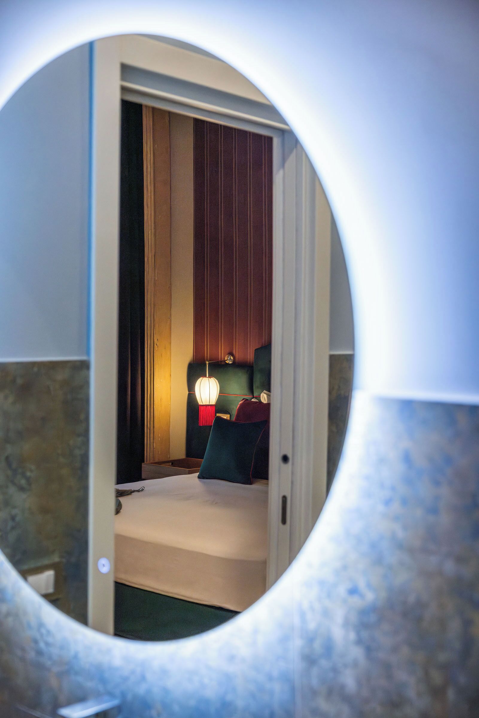 Espejo circular que refleja una habitación con una cama, una mesita de noche y una lámpara.