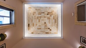 Une lucarne carrée ornée d’un motif en mosaïque, encadrée d’une bordure blanche, dans une pièce aux murs unis et dotée d’une fenêtre.