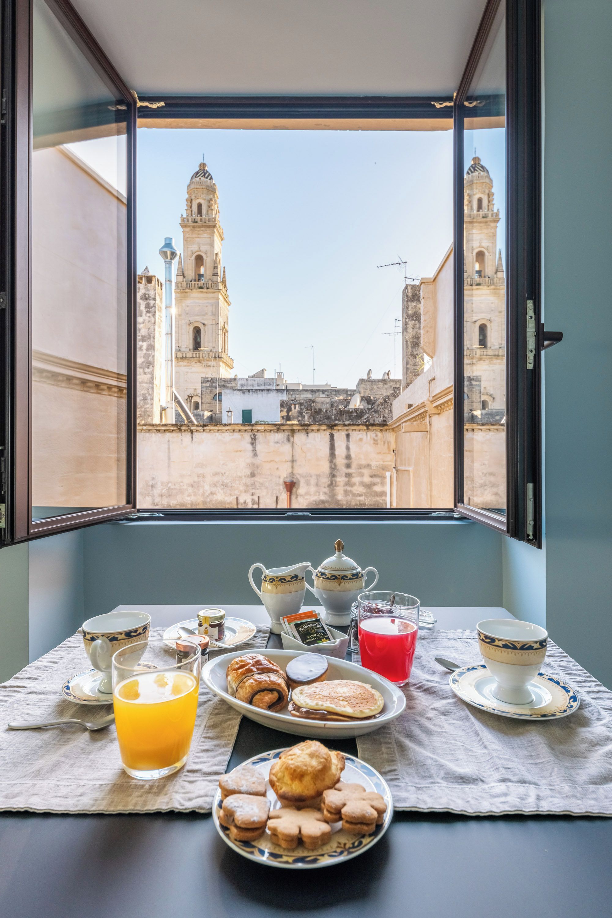 Ein Frühstückstisch mit Gebäck, einer Tasse Tee und einem Glas Orangensaft mit Blick auf historische Gebäude und Türme.