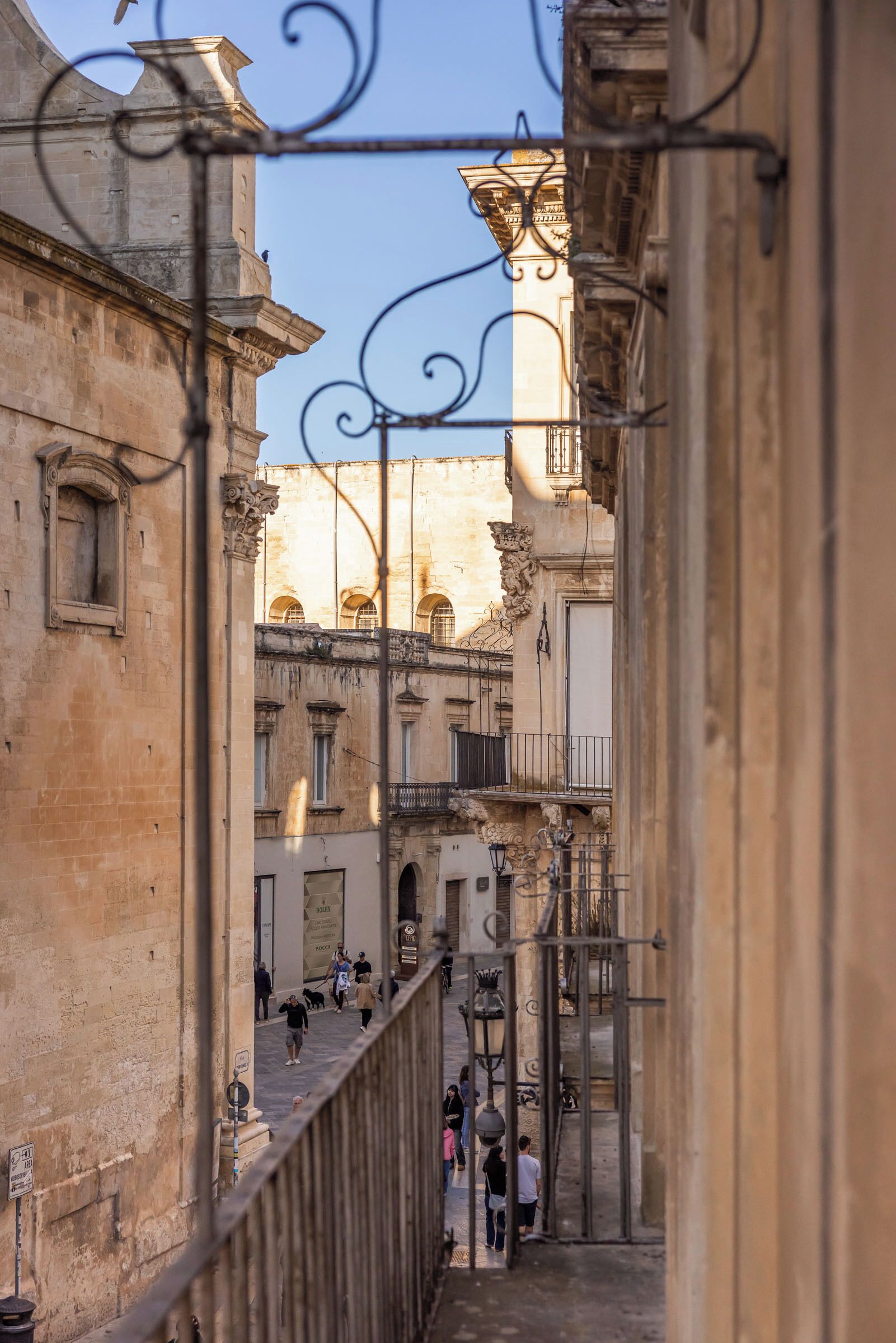 A narrow street with historic buildings, a metal railing, and people walking.