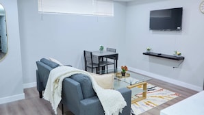 A modern living room with a sofa, a glass coffee table, a dining area with chairs, a TV mounted on the wall, and a small plant on a shelf. - Modern Basement Suite w/ Private Entrance (Winnipeg)