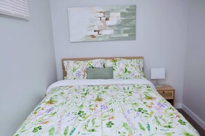 A bedroom with a floral bedspread, a wooden headboard, a bedside lamp, and a wall-mounted abstract painting. - Modern Basement Suite w/ Private Entrance (Winnipeg)