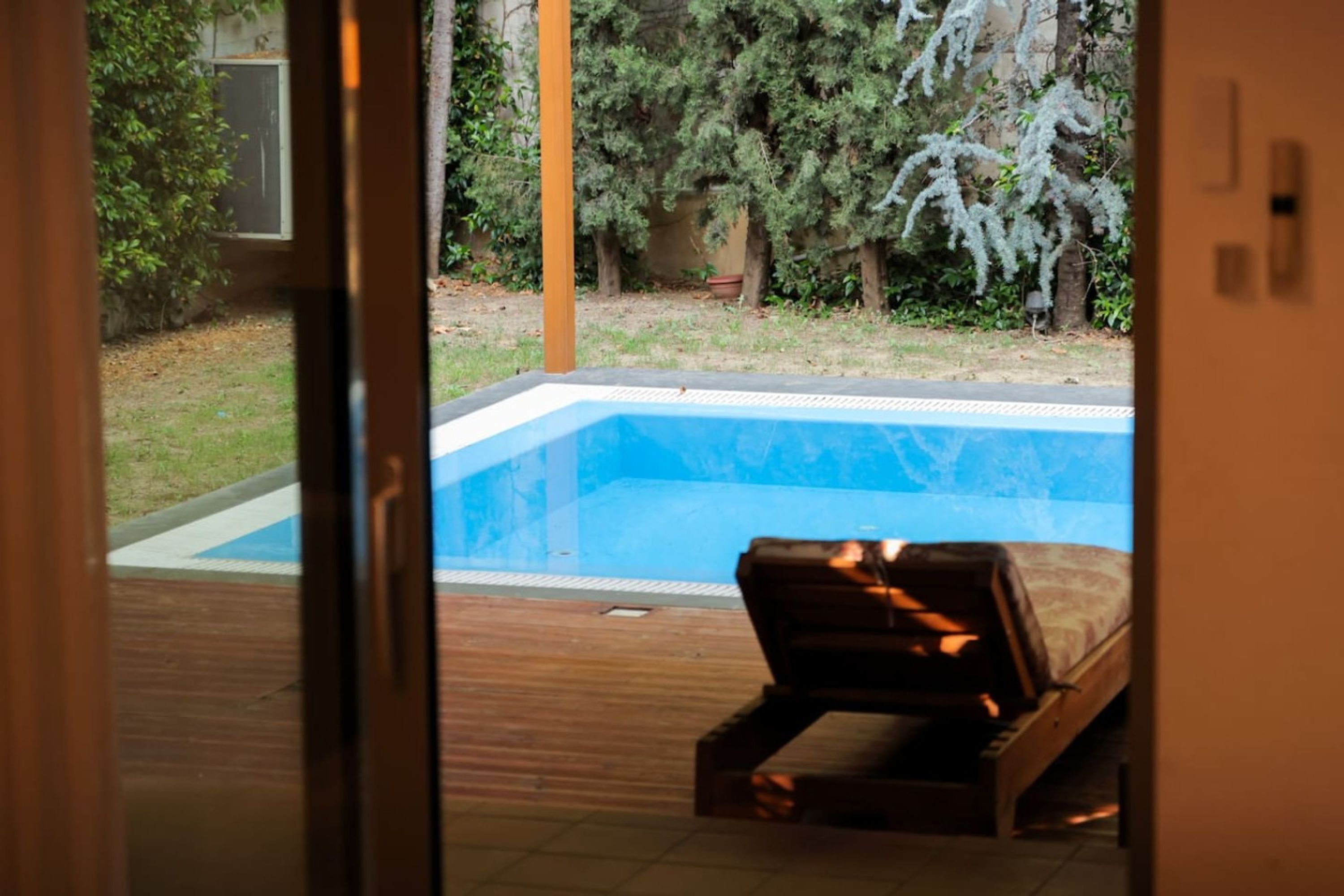 A poolside view with a wooden deck, a lounge chair, and a view of a backyard with trees and a swimming pool.