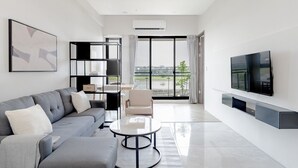 Uma sala de estar moderna com um sofá cinza, uma mesa de centro, uma TV montada na parede e uma janela grande com vista para o exterior.