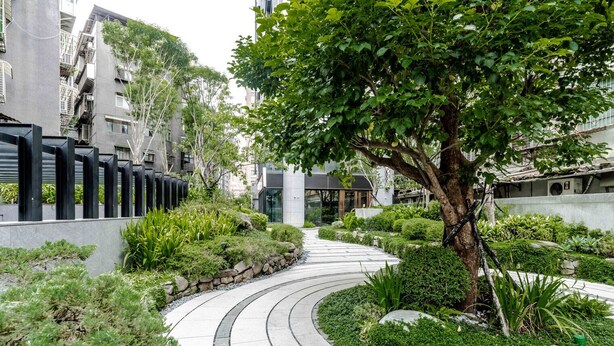 Un camí corbat en una zona enjardinada amb un arbre, vegetació i edificis moderns al fons.