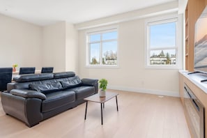A modern living room with a black leather sofa, a small wooden coffee table, and two large windows offering a view of the city. - Newly Built 3BR Townhome | Rooftop Patio & Garage (Langley)