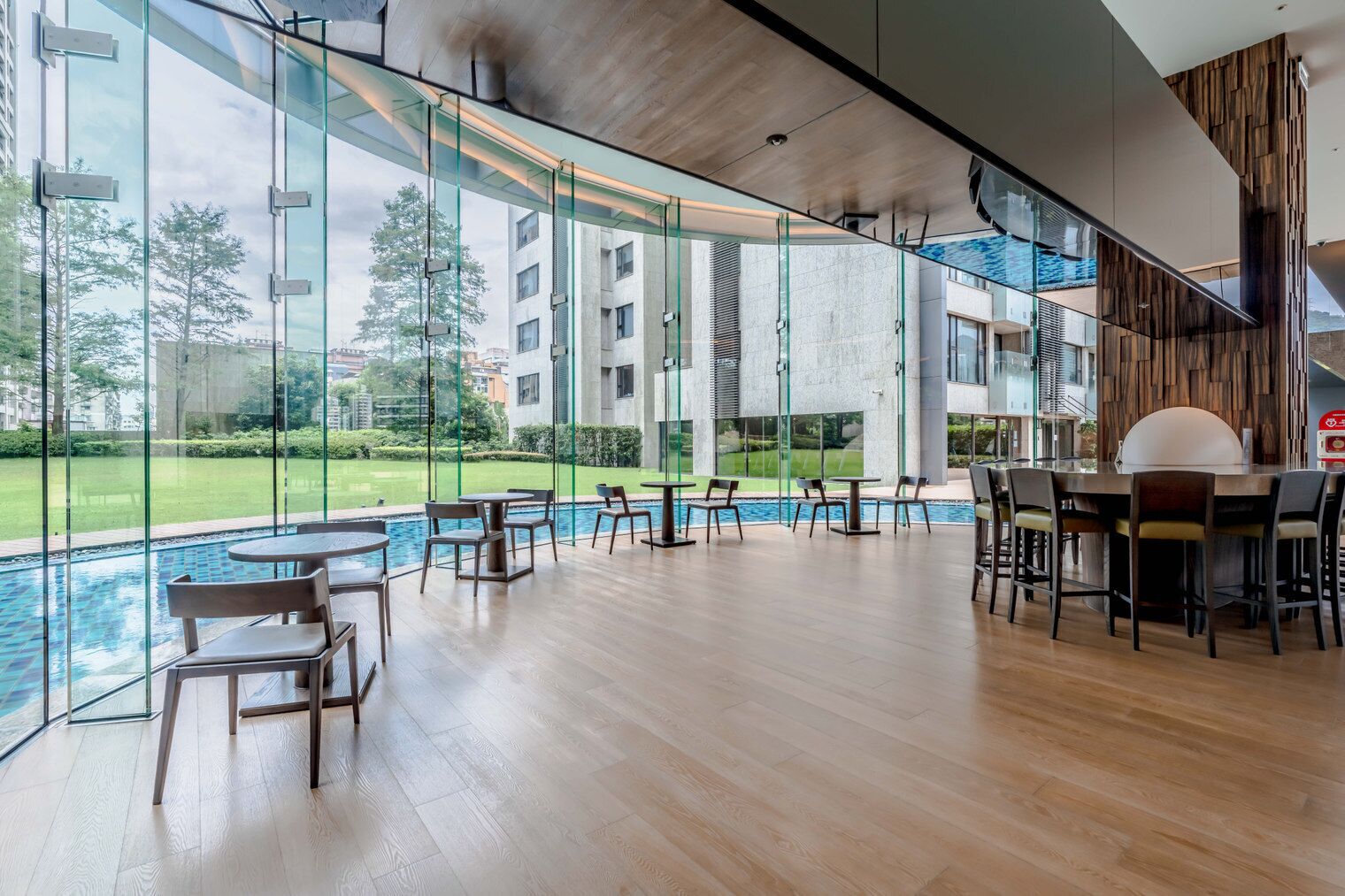 A modern indoor pool area with glass walls, wooden flooring, and multiple seating arrangements.
