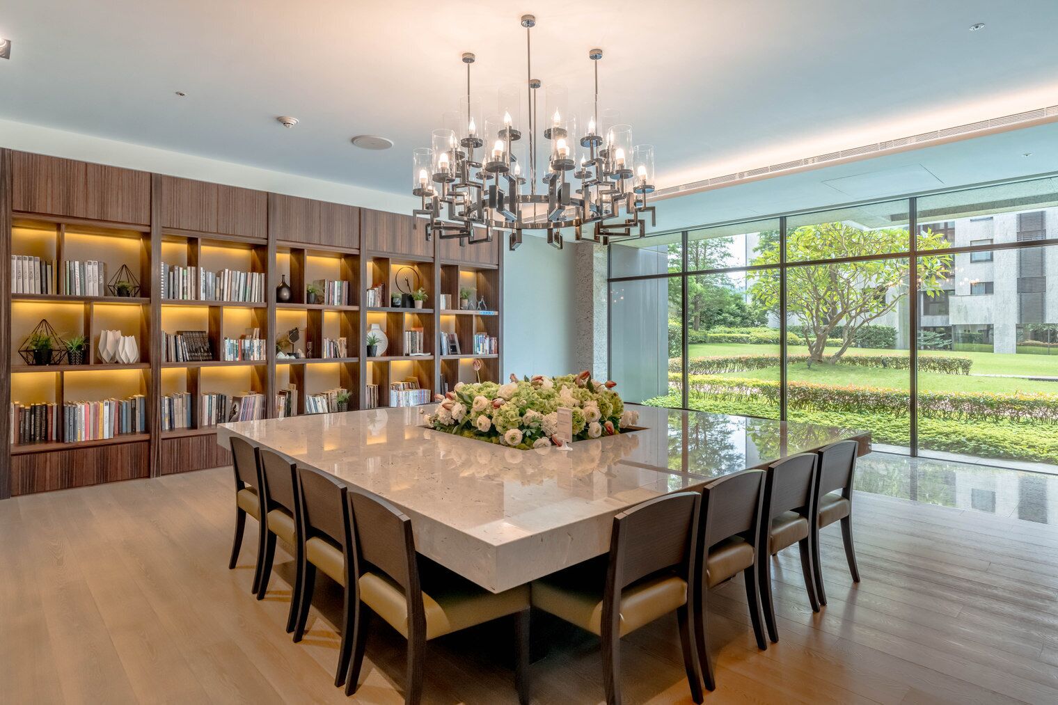 A modern kitchen with a large island, built-in bookshelves, and a view of a garden through large windows.