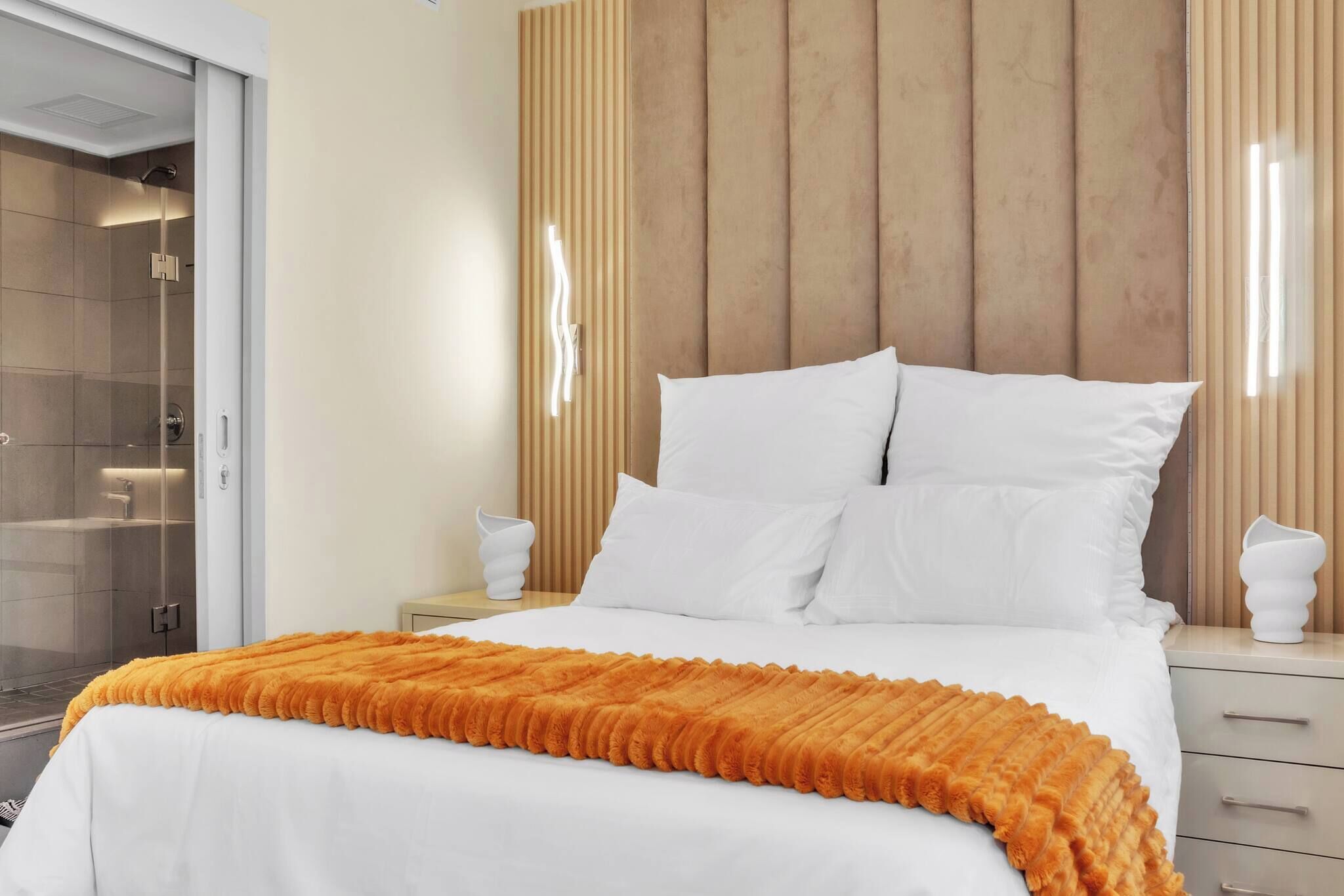 Un dormitorio moderno con una cama, almohadas blancas, un cabecero de madera, una mesita de noche y una ducha con cerramiento de vidrio.