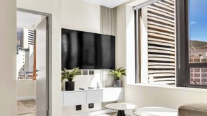 A modern living room with a flat-screen TV, a white console, a round table, and a large window with a view of buildings. - Softstay, Foreshore Cape Town CBD (Cape Town)