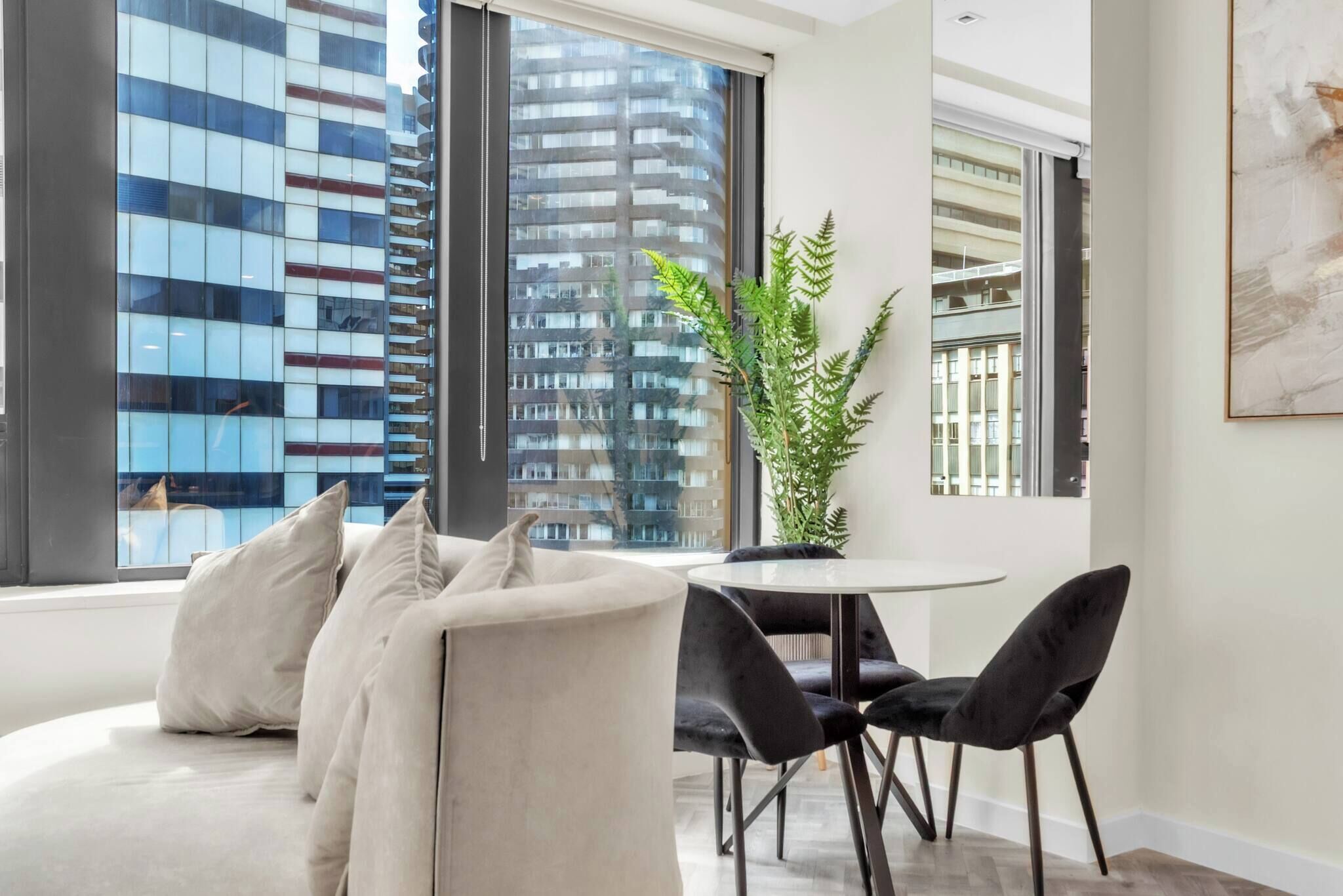A modern living room with a sofa, a dining table with chairs, a potted plant, and large windows offering a view of high-rise buildings.