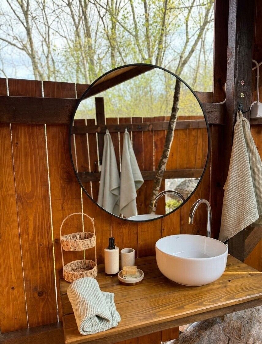 A wooden cabin bathroom with a round mirror, a white sink, a hanging towel, and a small shelf with toiletries.