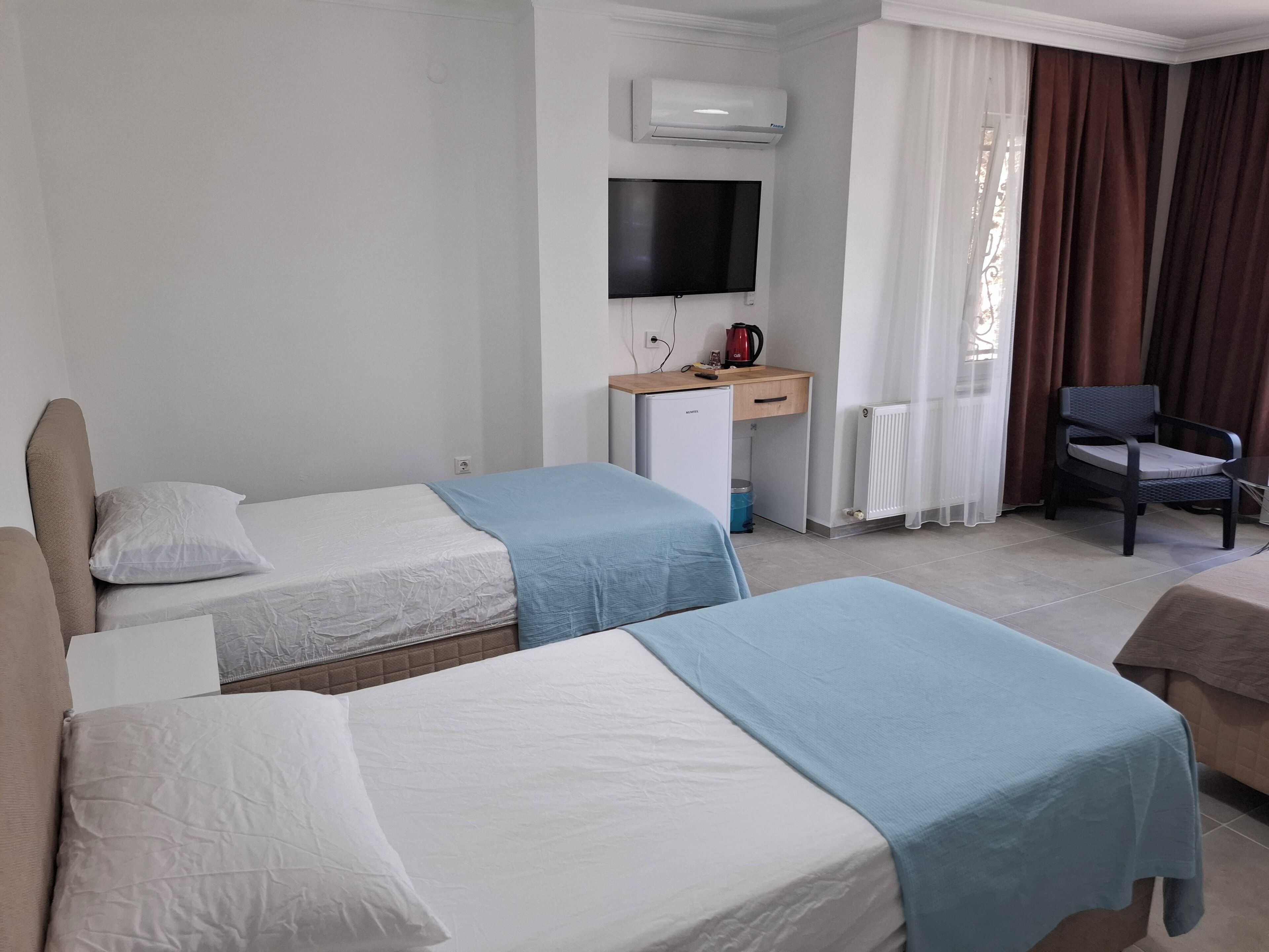 Ein Hotelzimmer mit zwei Einzelbetten, einem Schreibtisch mit Fernseher, einem Sessel und einem Fenster mit Vorhängen.