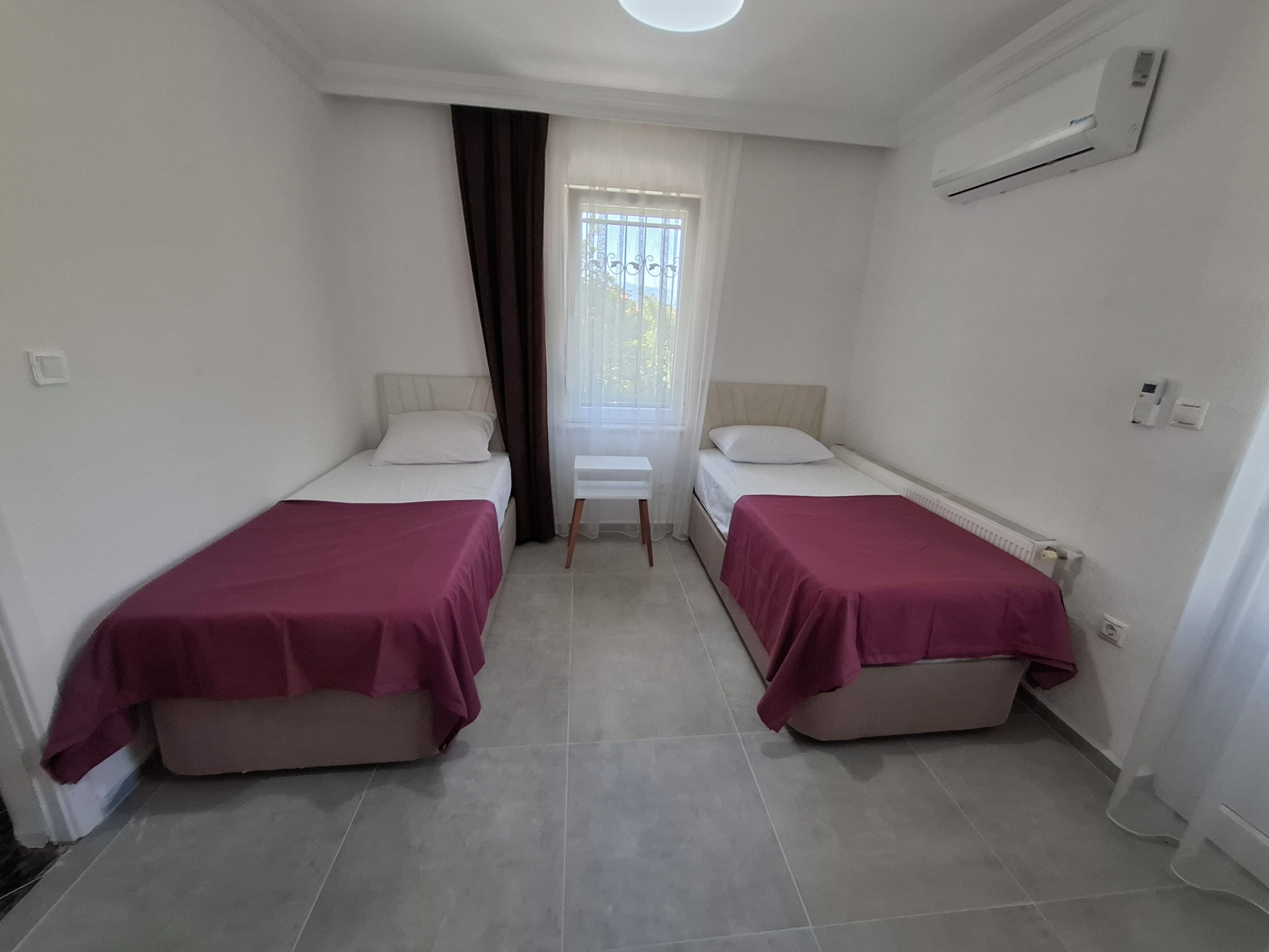 A small, clean room with two single beds, a window with curtains, a small table, and an air conditioning unit.
