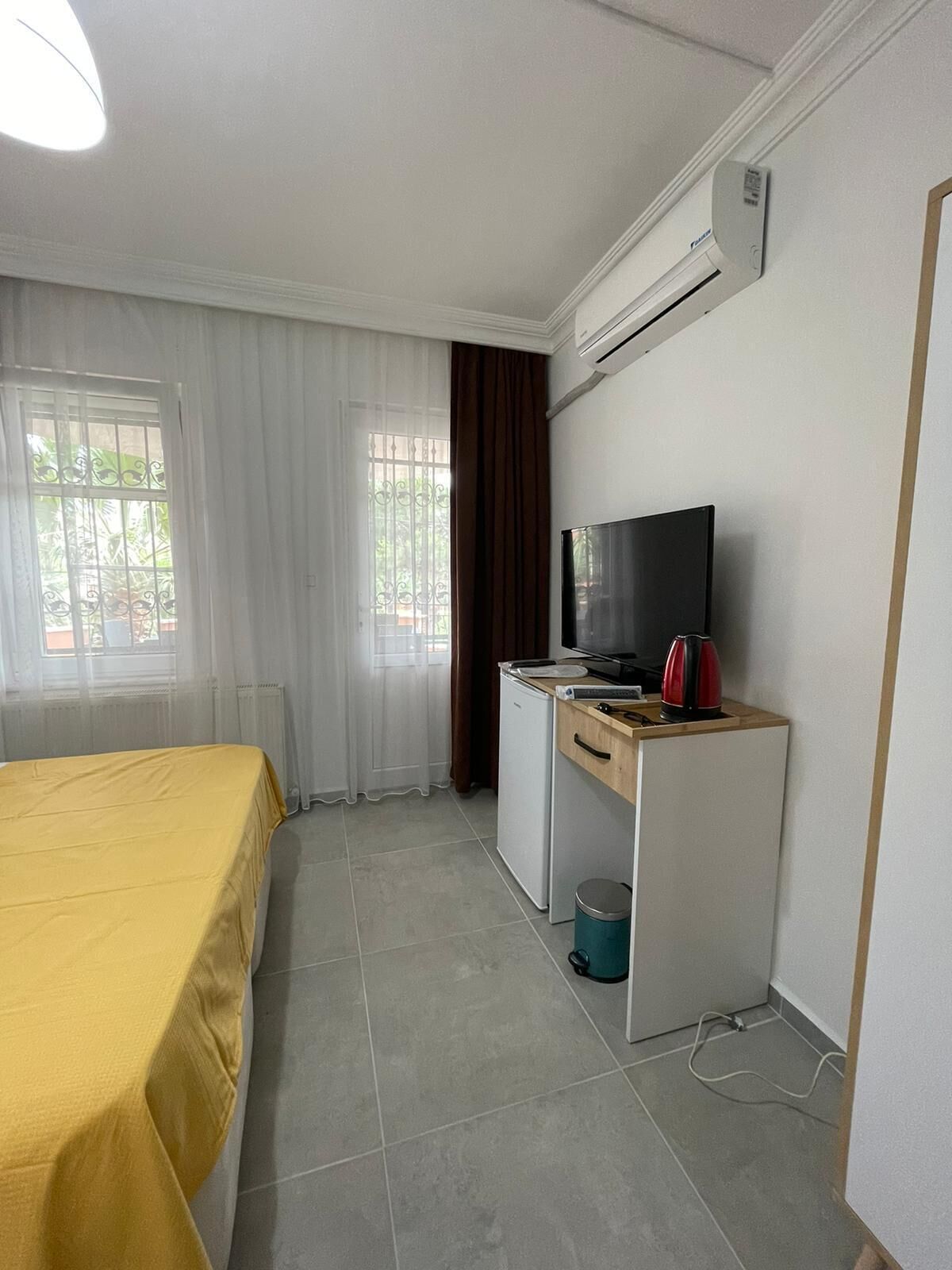 A hotel room with a bed, a desk with a TV, a small fridge, a kettle, and a window with curtains.