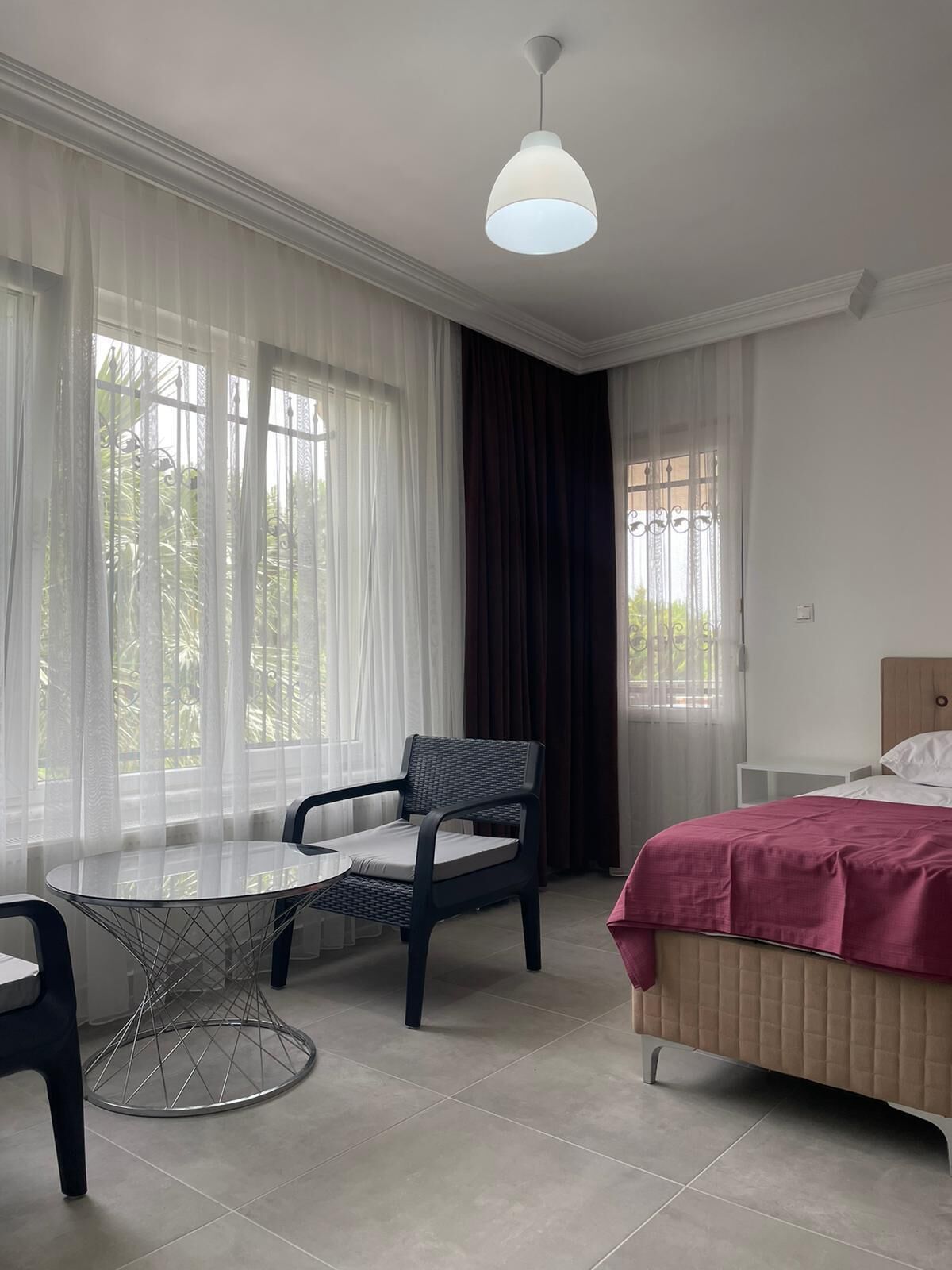 Habitación de hotel con una cama, una mesa redonda con una silla, una lámpara colgante y ventanas amplias con cortinas traslúcidas.