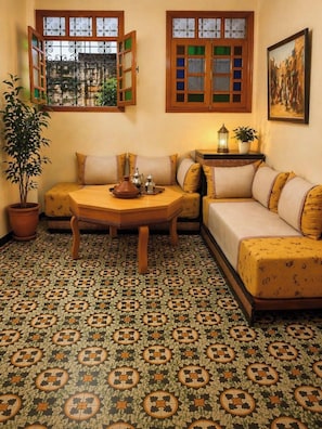 Ein Zimmer mit einem gemusterten Boden, einem Sofa, einem Tisch mit einer Vase und zwei Fenstern mit geöffneten Fensterläden.