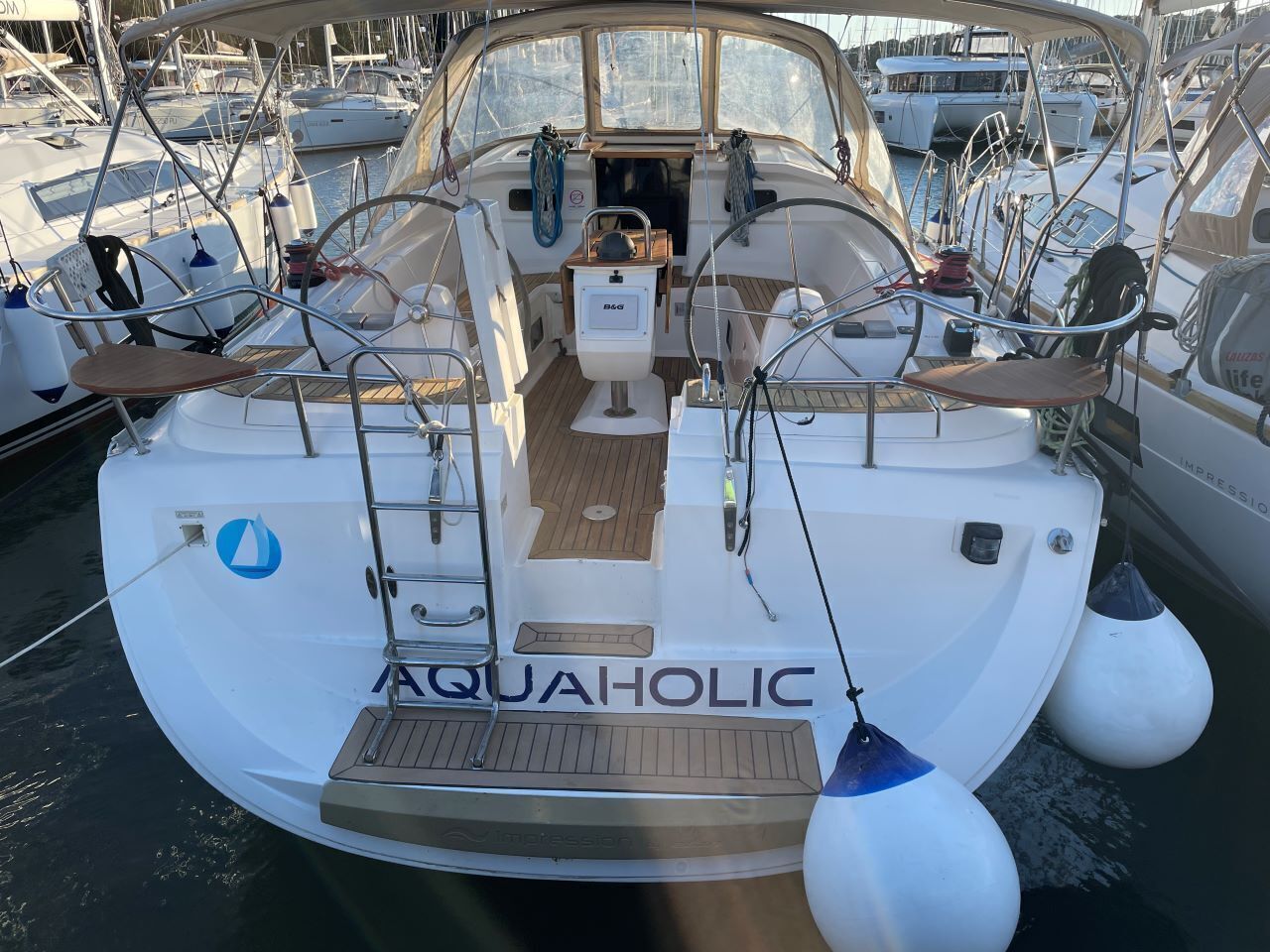 Ein Boot namens AQUAHOLIC liegt in einer Marina mit Holzterrasse, Steuerrad und einem kleinen Kabinenbereich festgemacht.