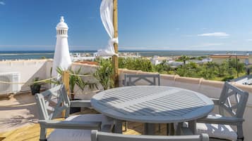 A rooftop terrace with a round table and chairs, a white lamp, and a view of the sea and buildings.