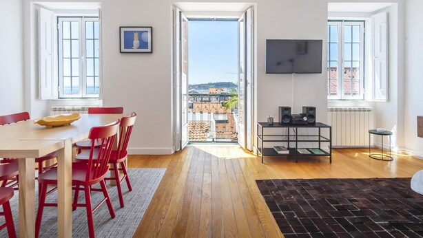 A spacious room with a wooden floor, a dining table with red chairs, a black TV on the wall, and a view of buildings through a door. - BI1 in Lisboa Ciudad (Lisbon)