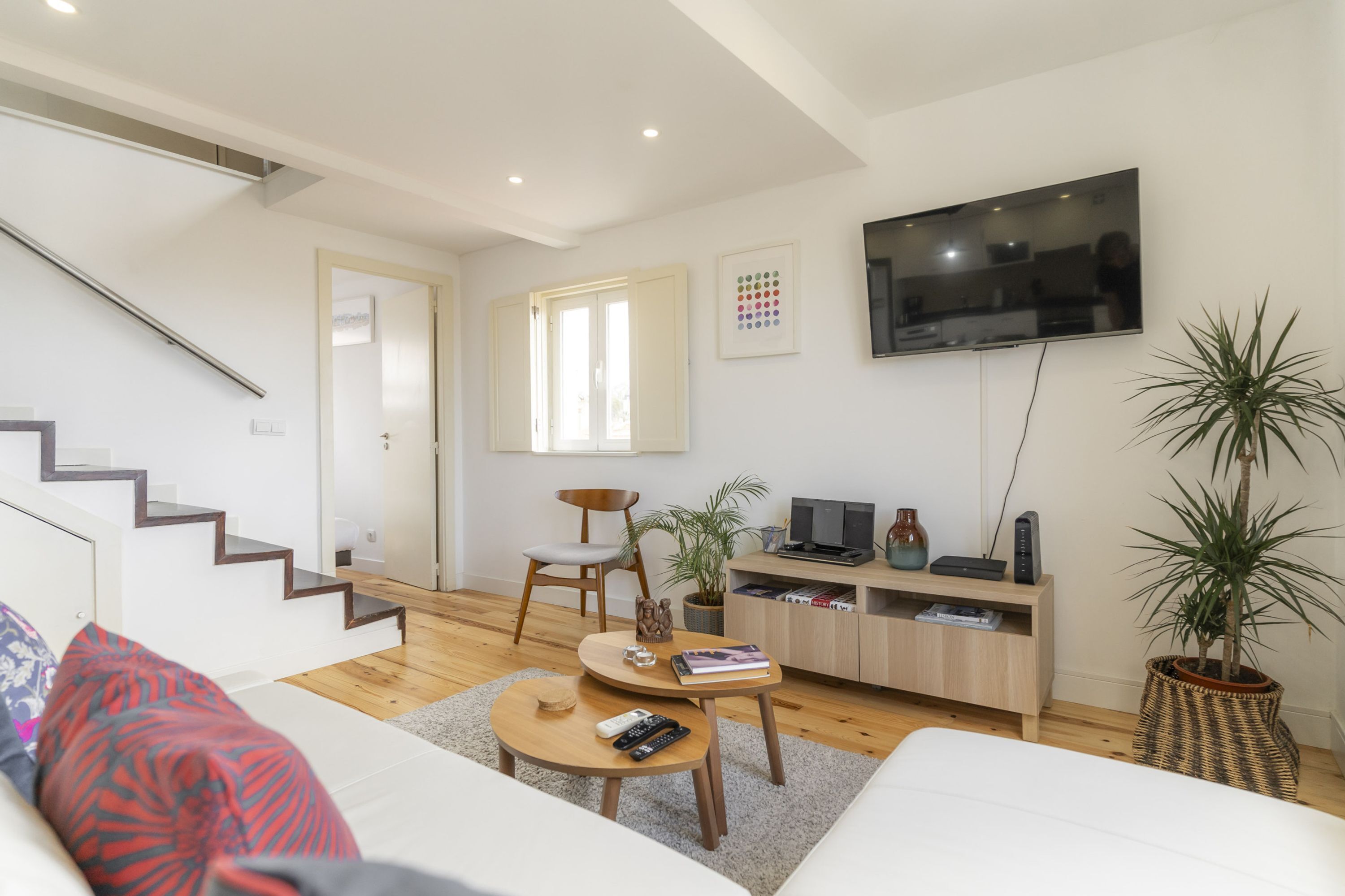 A modern living room with a staircase, a flat-screen TV, a wooden coffee table, a sofa, and a potted plant.
