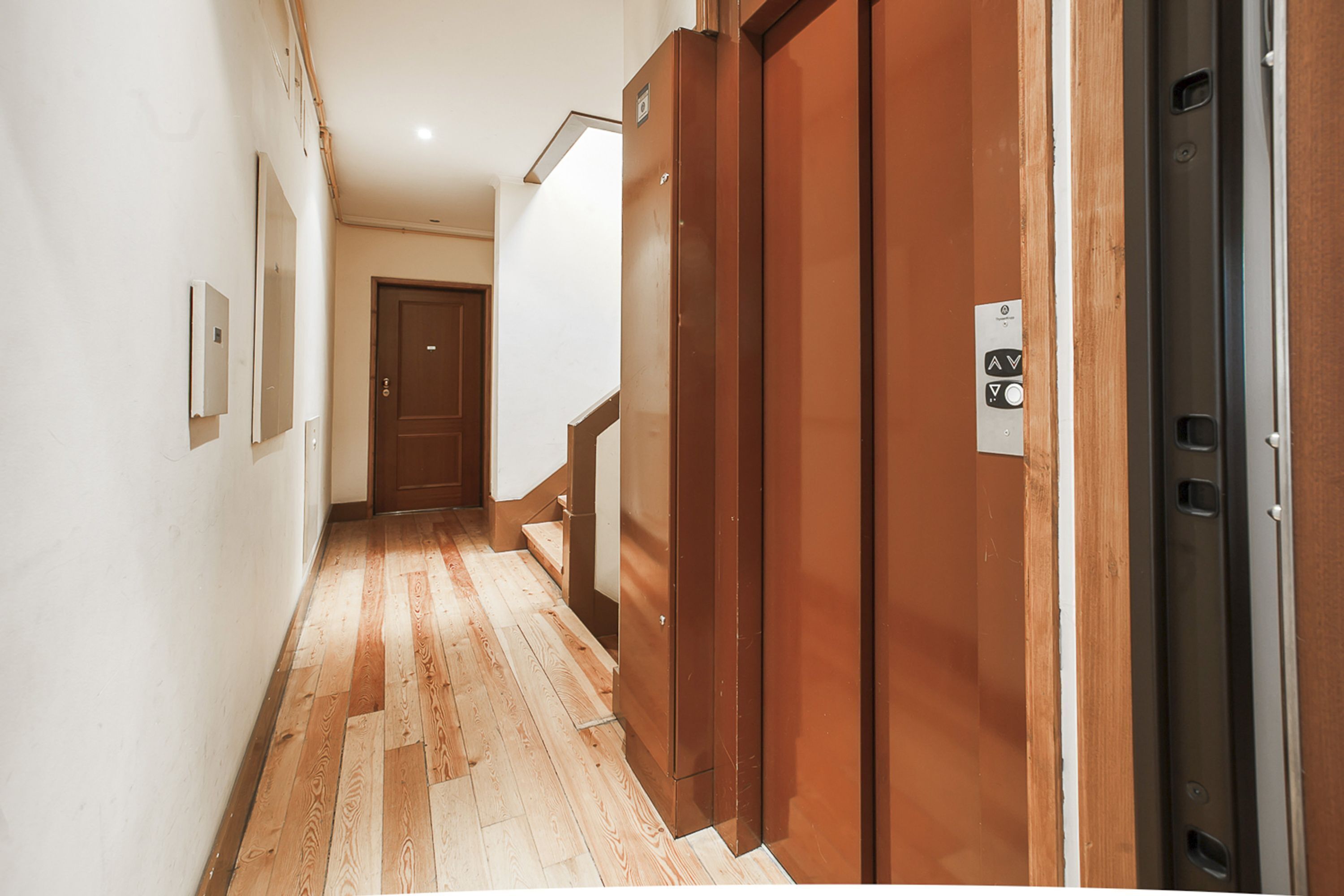 A hallway with wooden flooring, a door, and an elevator.