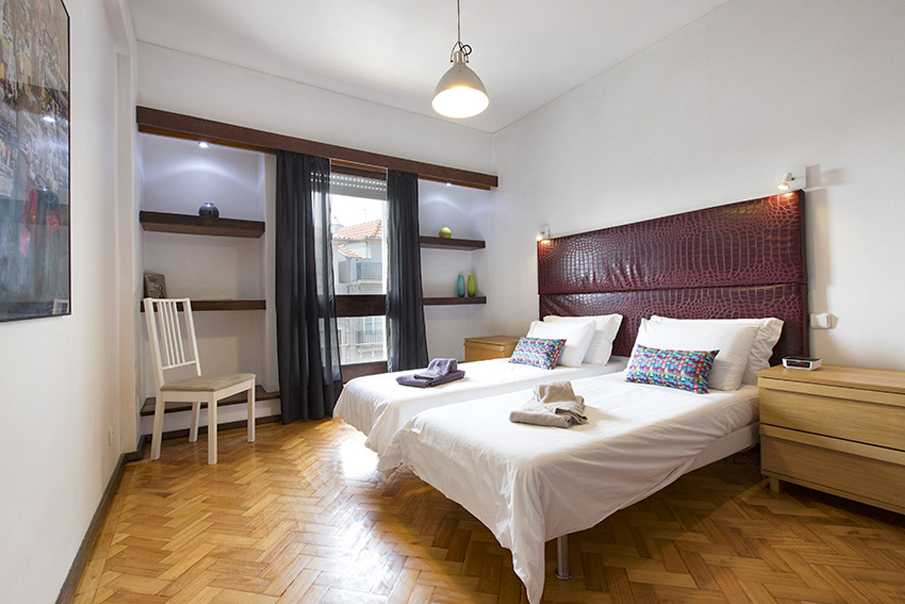A bedroom with two beds, a chair, a bedside table, a nightstand, and a window with curtains.