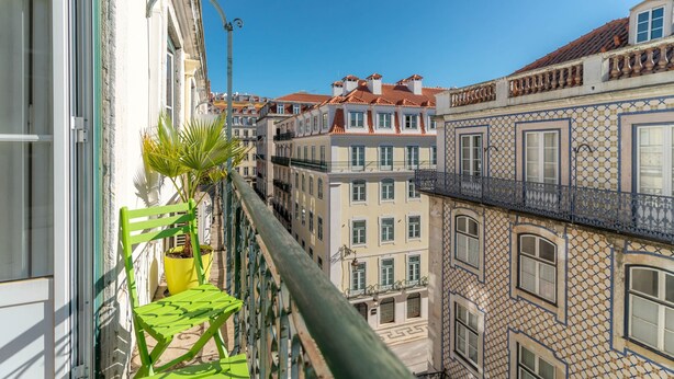 Uma varanda com cadeira e planta em vaso, com vista para uma fileira de edifícios com arquitetura tradicional e varandas.