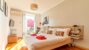 Quarto com cama grande, mesa de cabeceira, cadeira, janela e televisão fixada na parede.