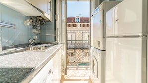 Cozinha pequena com pia, máquina de lavar e vista para uma varanda com telhado em azulejos.