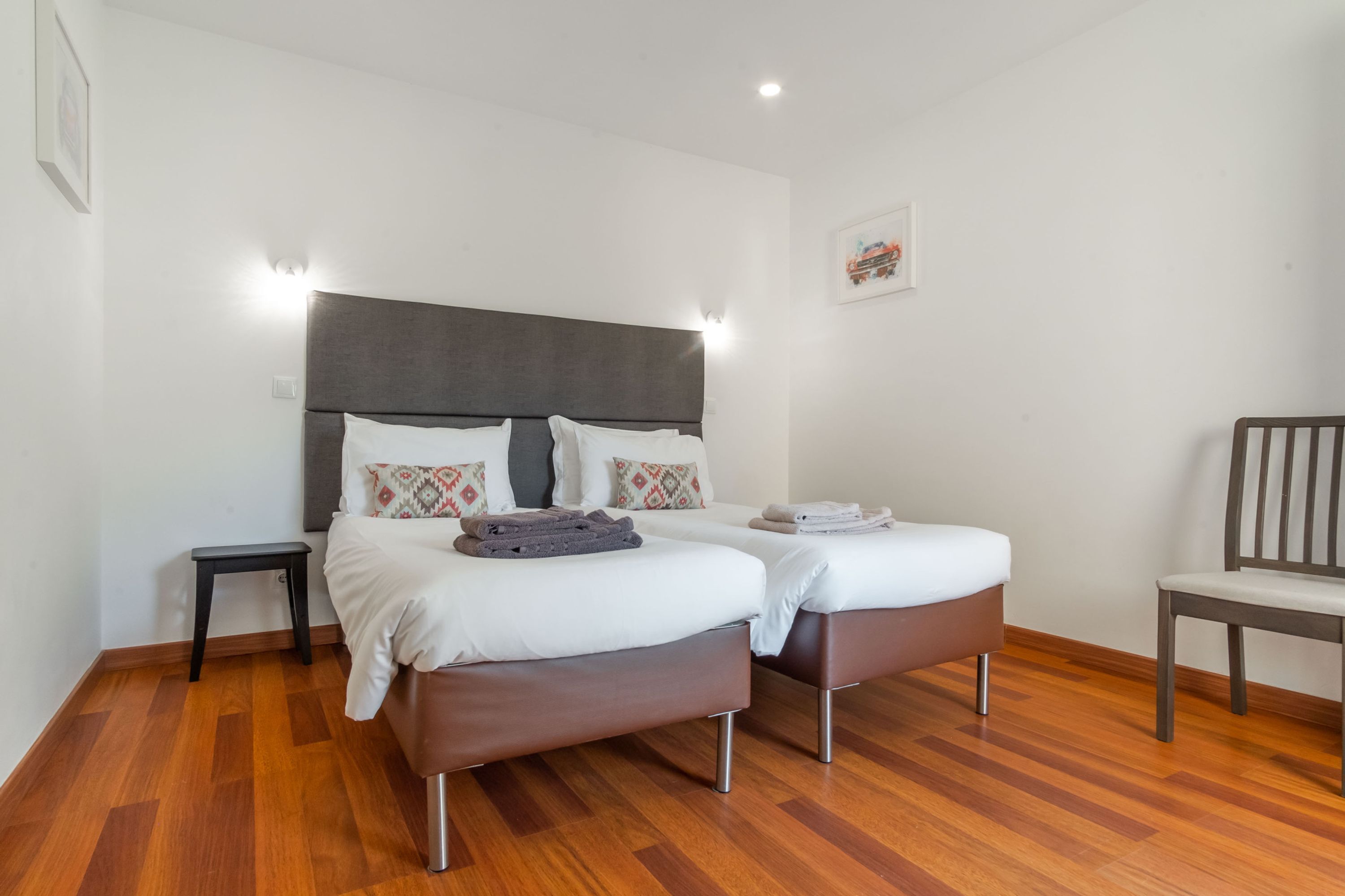 A double bed with a grey headboard, two bedside tables, a chair, and a wooden floor.