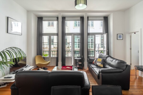 A modern living room with a black leather sofa, a yellow chair, a flat-screen TV, and large windows with curtains. - FI in Lisboa Ciudad With 2 Bedrooms and 1 Bathrooms (Lisbon)