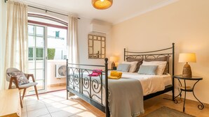 Un dormitorio con cama, una silla, una mesa, una lámpara y una ventana.