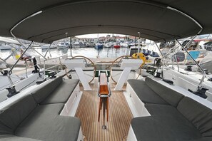 Convés de barco com assentos, leme e vista para um porto com vários barcos.