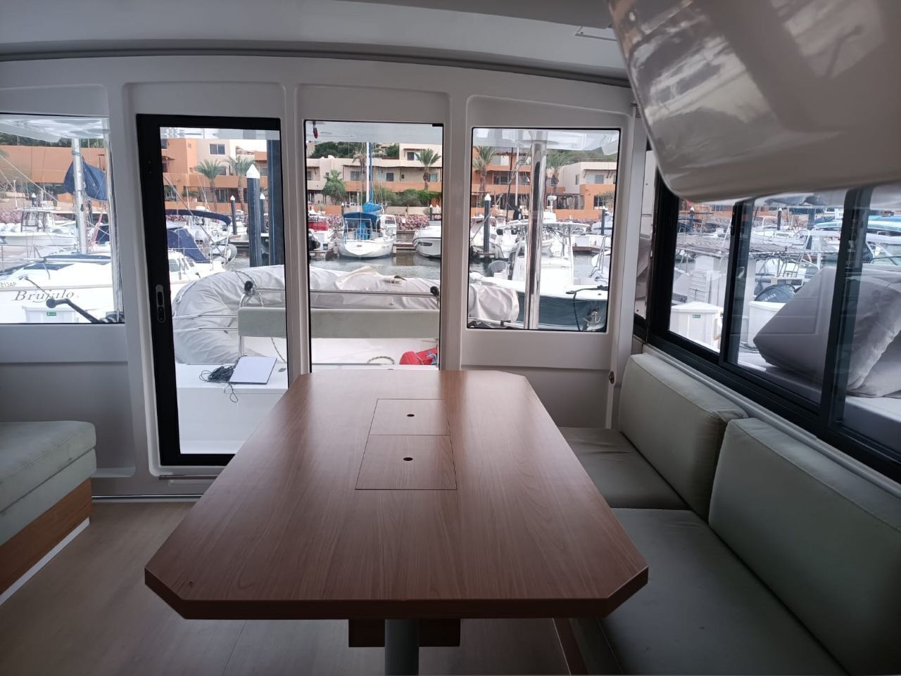 Interior de barco com mesa de madeira, assentos acolchoados e vista para uma marina com barcos e edifícios.