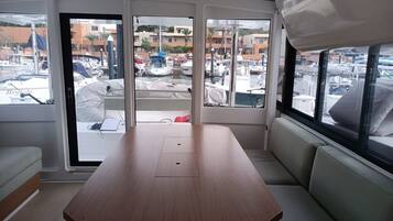 A boat interior with a wooden table, cushioned seating, and a view of a marina with boats and buildings.