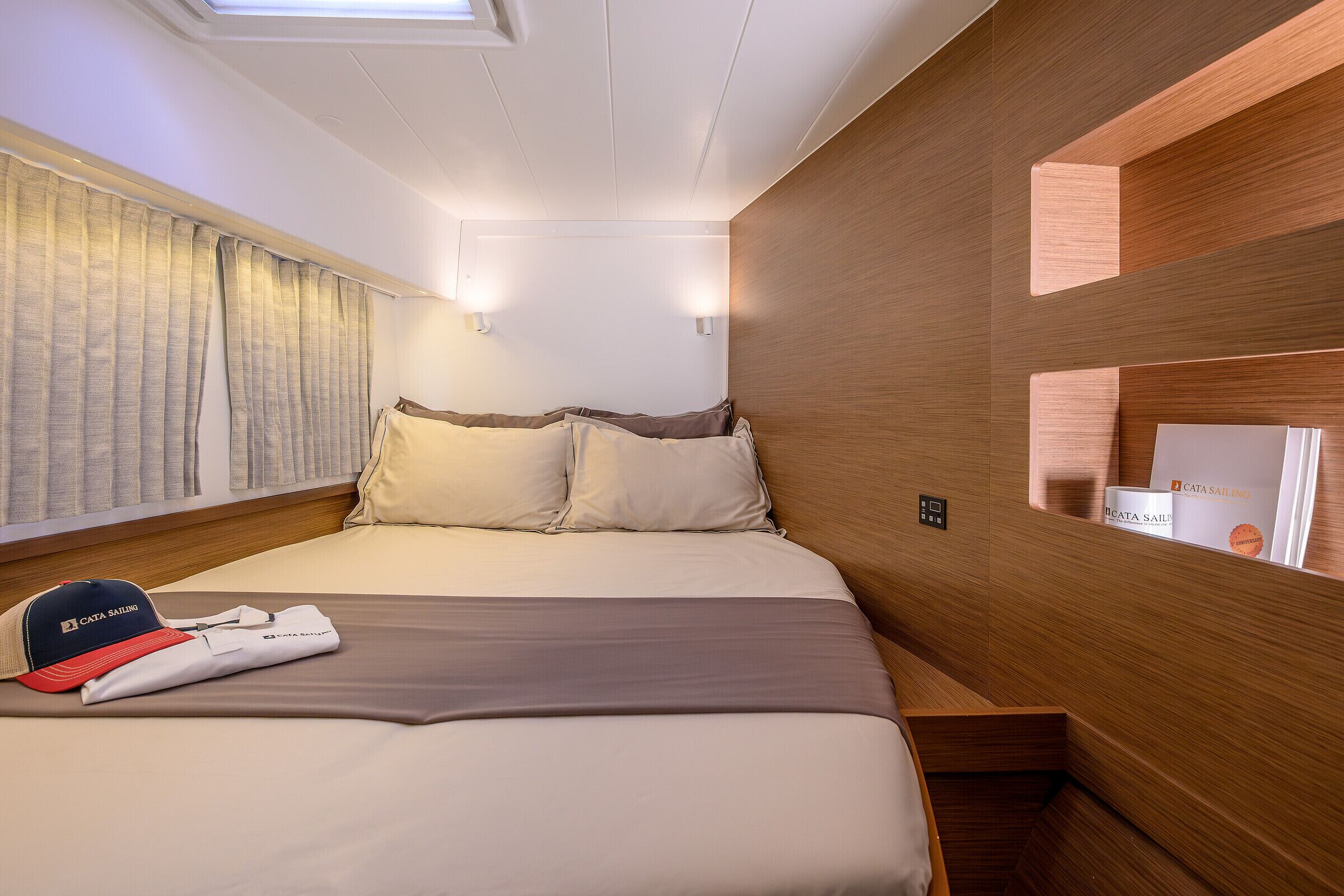 A neatly made bed with pillows, a folded towel, and a cap on the bed. There are two wall-mounted lights and a window with curtains.