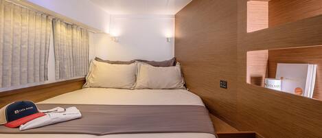 A neatly made bed with pillows, a folded towel, and a cap on the bed. There are two wall-mounted lights and a window with curtains.