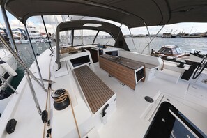 Cubierta de barco con mesa de madera, una consola blanca y vistas a otras embarcaciones y un puerto deportivo.