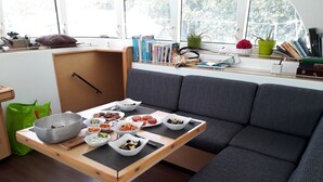 Un salon moderne avec un canapé gris, une table en bois sur laquelle sont disposés des bols remplis de nourriture, et une fenêtre ornée de livres et de plantes.