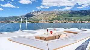 Een scheepsdek met een tafel, lounge stoelen en uitzicht op een kustplaatsje en bergen.