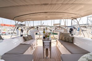 Eine überdachte Terrasse mit Sitzgelegenheiten, einem Tisch mit einer Flasche und Blumen sowie Blick auf andere Boote.
