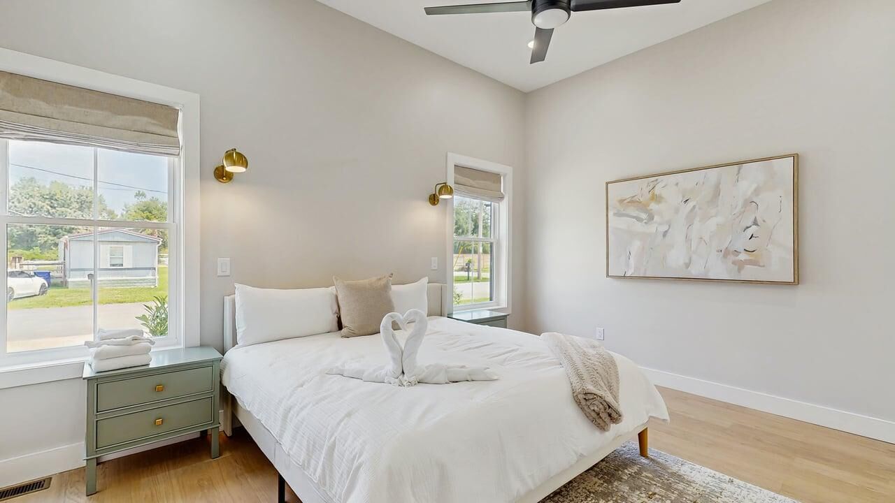Un dormitorio con cama, mesitas de noche, ventilador de techo, ventana con vistas y un cuadro en la pared.