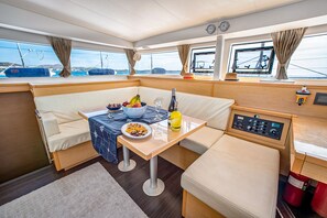 A boat's interior with a dining area, cushioned seating, a table with food and drinks, and a control panel. - Sun-Kissed Sailing Adventure in Islas Sarónicas (Alimos)